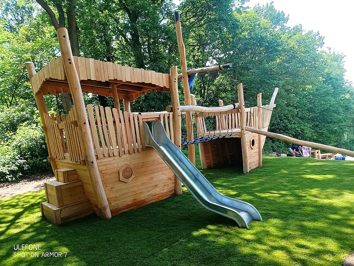 Großes Holz-Spielschiff mit Edelstahlrutsche, Kletternetz und Steuerrad auf grüner Wiese – umgeben von Bäumen im Park