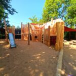 „Großes Holzspielgerät namens Piratenbollwerk mit Rutsche, Netzen und Kletterelementen auf Sandfläche unter schattenspendenden Bäumen.“