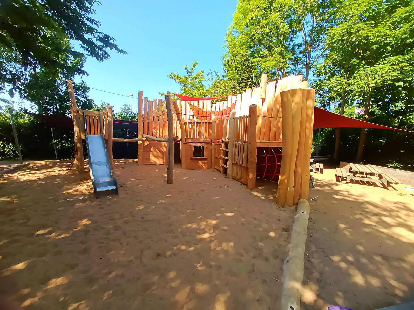 „Großes Holzspielgerät namens Piratenbollwerk mit Rutsche, Netzen und Kletterelementen auf Sandfläche unter schattenspendenden Bäumen.“