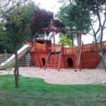 Großes Spielschiff aus Holz mit Edelstahlrutsche, Kletterelementen und Sandfläche – eingebettet in einen umzäunten, grünen Spielbereich