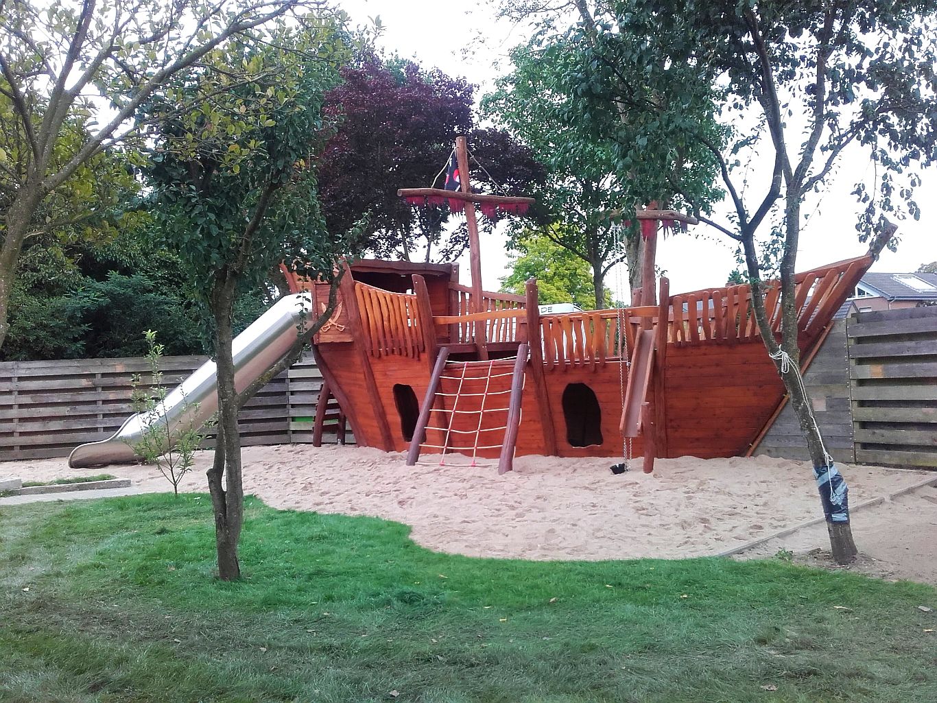 Großes Spielschiff aus Holz mit Edelstahlrutsche, Kletterelementen und Sandfläche – eingebettet in einen umzäunten, grünen Spielbereich