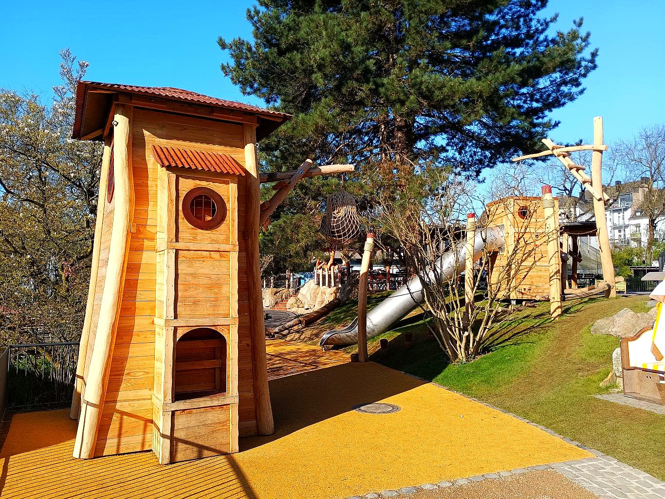 „Spielbereich im Zoo im Stil eines Hafens mit hölzernen Türmen, Seilröhre und Edelstahlrutsche – eingebettet in eine naturnahe Hügellandschaft.“