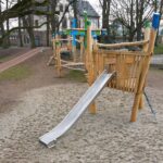 Langes Holz-Spielschiff mit Edelstahlrutsche, Kletterelementen und Hängebrücken – Spielplatz mit Naturmaterialien neben einer Schule