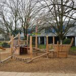 „Holz-Spielschiff auf Spielplatz mit bunten Kletterelementen, Balancierstrecken und Seilbrücke – in einem Schulhof mit Bäumen.“
