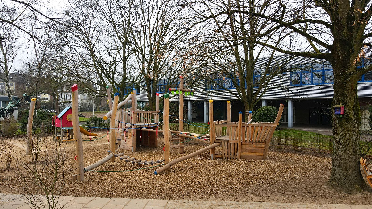 „Holz-Spielschiff auf Spielplatz mit bunten Kletterelementen, Balancierstrecken und Seilbrücke – in einem Schulhof mit Bäumen.“