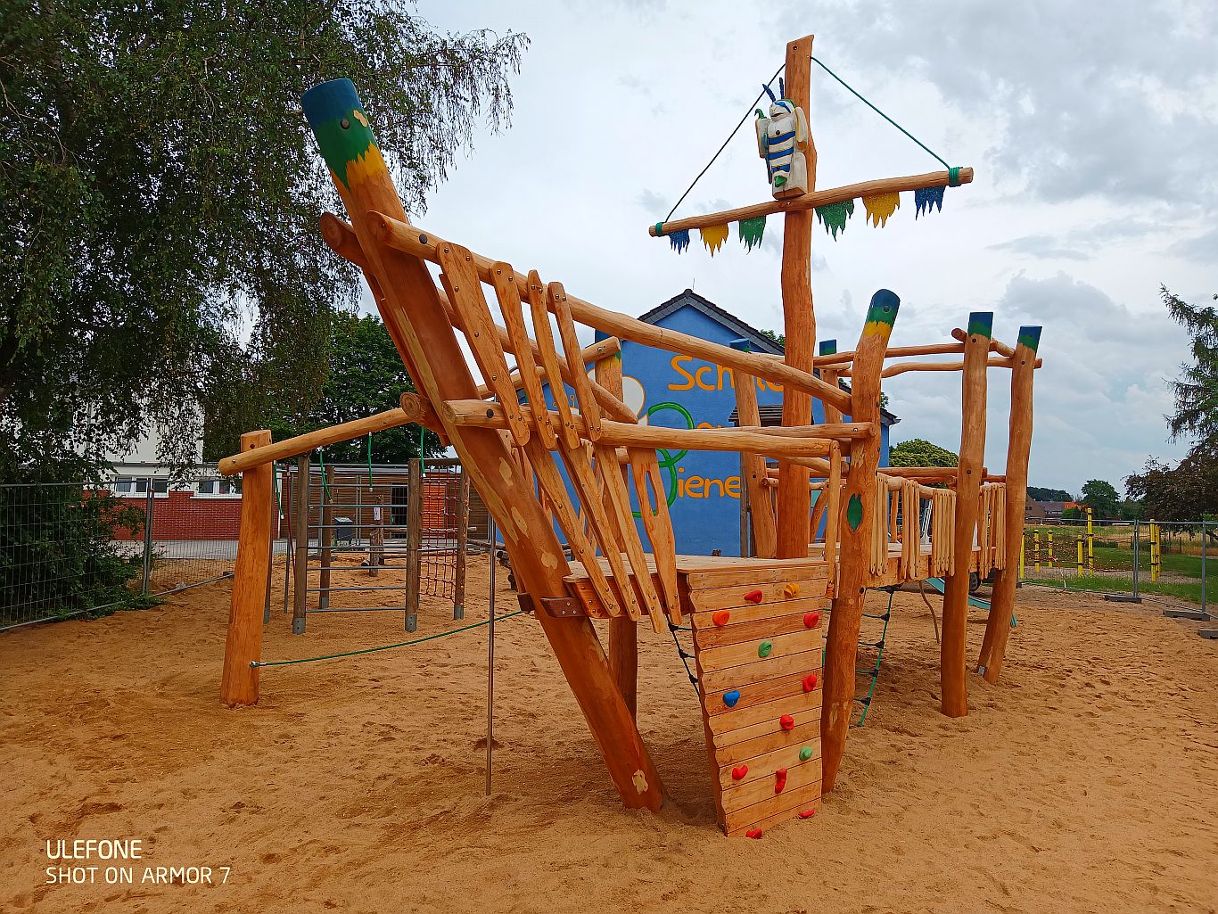Holz-Spielschiff auf Sandfläche mit buntem Kletteraufstieg, Masten und Seilen – vor einer blau gestrichenen Schulwand mit Schriftzug