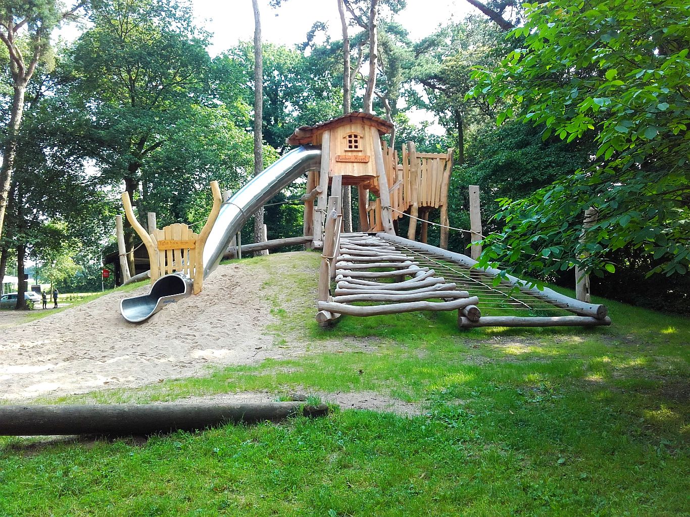 Naturnaher Spielplatz mit Holz-Kletterhaus, großer Rutsche, Kletterrampe und Sandfläche – eingebettet im Waldgebiet