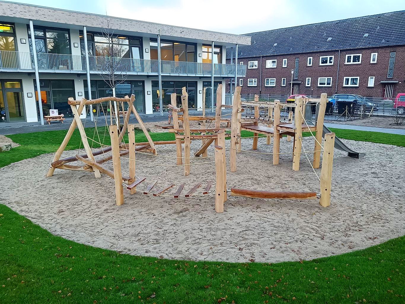 Ein modern gestalteter Spielplatz mit Spielgeräten aus Robinienholz, darunter Kletterstrukturen, Schaukeln und Balancierbalken, auf einem Sandboden. Gelegen in einer Wohnanlage mit Mehrfamilienhäusern im Hintergrund.