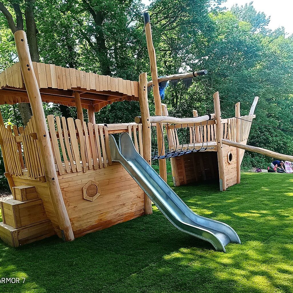 Holz-Spielschiff mit Rutsche und Seilbrücke Spielplatz Robinie Großes Holz-Spielschiff mit Edelstahlrutsche, Kletternetz und Steuerrad auf grüner Wiese – umgeben von Bäumen im Park