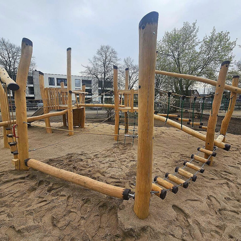 Kletteranlage Spielplatz Kindergarten Ein rustikaler Spielplatz mit robusten Holzklettergeräten auf einem Sandboden, umgeben von urbaner Landschaft. Die Anlage bietet verschiedene Klettermöglichkeiten und Balancierstrecken, ideal für die Förderung von Motorik und Gleichgewicht bei Kindern.
