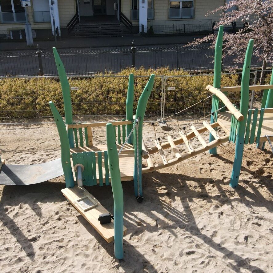 kletterspielplatz-aus-holz Ein urbaner Spielplatz mit Klettergerüsten und Rutschen in leuchtendem Grün, positioniert auf einem sandigen Untergrund. Die Spielgeräte aus Holz sind von Wohngebäuden umgeben und an einer Straße mit geparkten Autos gelegen. Blühende Bäume im Hintergrund verleihen der Szene eine frühlingshafte Atmosphäre.