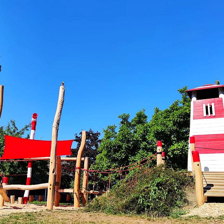 Maritimer-Spielplatz-Leuchtturm-Holz Maritimer Spielplatz mit rotem Sonnensegel, Leuchtturm-Kletterturm und Holzbrücke – umgeben von Bäumen und Natur