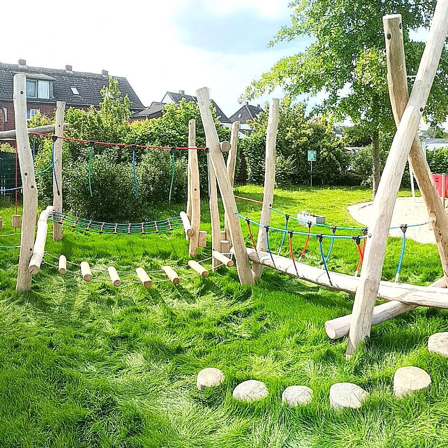 balancierparcours-kita Ein farbenfroher Balancierparcours aus Holz in einem parkähnlichen Kinderspielplatz, umgeben von grünem Gras und Bäumen. Die Anlage beinhaltet Schaukeln, Seilbrücken und balancierende Baumstämme, ideal für kindliche Koordinations- und Gleichgewichtsübungen. Im Hintergrund sind weitere Spielgeräte und Wohnhäuser sichtbar.