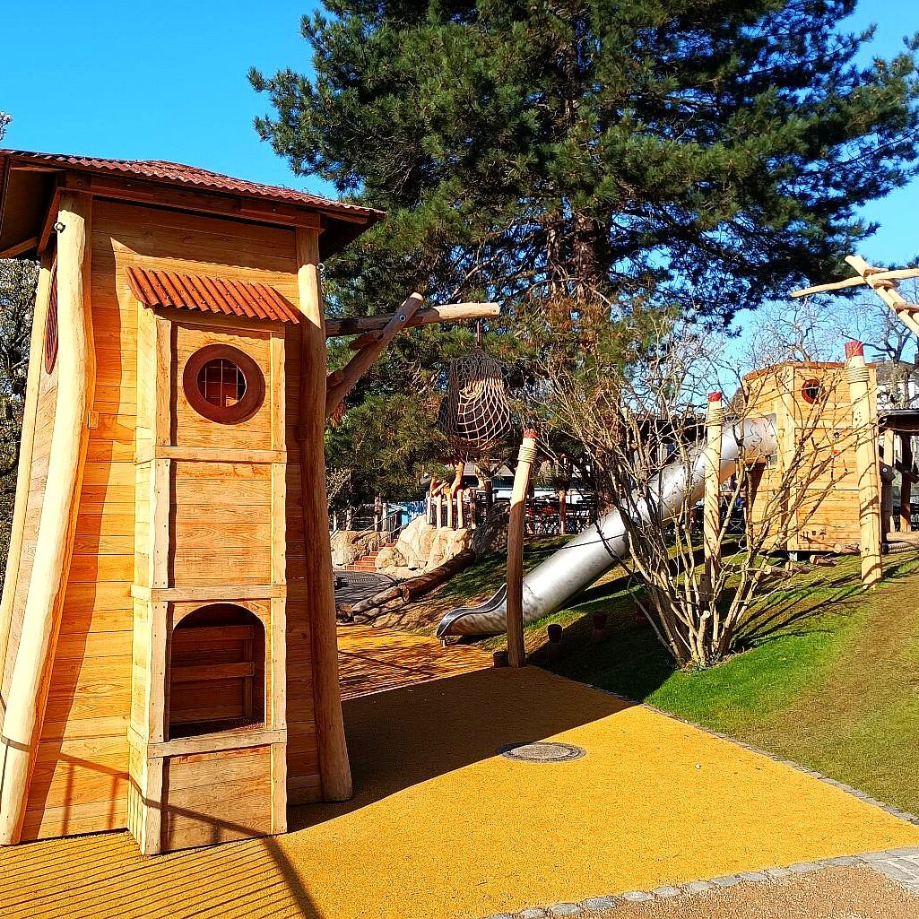 hafen-spielplatz-zoo-holzturm-rutsche „Spielbereich im Zoo im Stil eines Hafens mit hölzernen Türmen, Seilröhre und Edelstahlrutsche – eingebettet in eine naturnahe Hügellandschaft.“