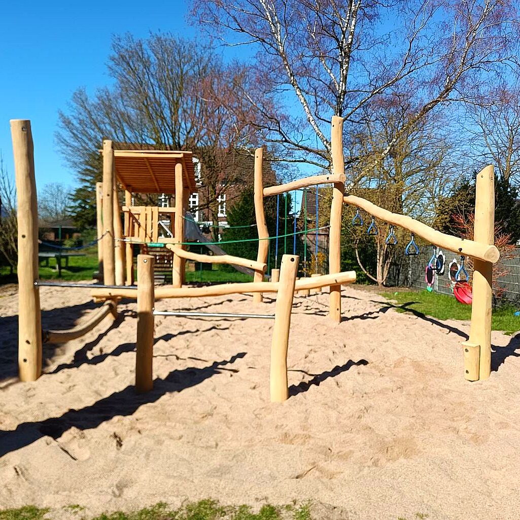 kletterparcours-kita-naturnah Ein sonniger Spielplatz mit einem naturnahen Kletterparcours aus Holz, umfasst verschiedene Klettermöglichkeiten wie Seile und Balken, gelegen auf einem Sandboden. Im Hintergrund sind Bäume und Wohngebäude sichtbar.