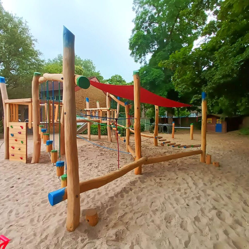 naturholz-spielplatz-kletteranlage Ein farbenfroher Spielplatz mit natürlichen Holzelementen und vielfältigen Spielgeräten auf einem Sandboden. Die Anlage umfasst eine Kletterstruktur mit Seilen, eine Rutsche, schwingende Brücken und eine Klettersprossenwand, unter einem rot schattierenden Sonnensegel. Umgeben von üppigem Grün bietet der Spielplatz einen ansprechenden Ort für Kinder, um in sicherer und naturnaher Umgebung zu spielen.
