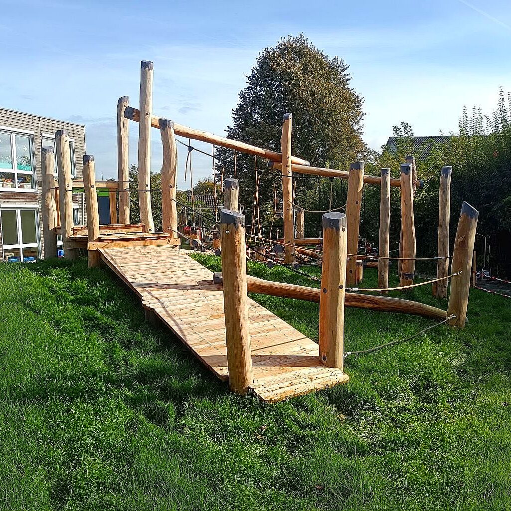 niedrigseilgarten-auf-hügel-kita Ein Niedrigseilgarten aus natürlichem Holz auf einem grünen Hügel neben einer Kindertagesstätte, konzipiert für Balance- und Kletteraktivitäten für Kinder.