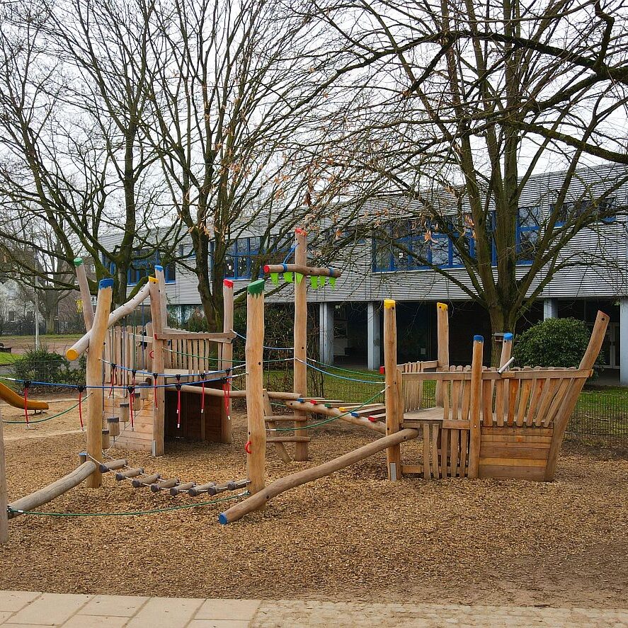 spielschiff-holz-kletterelemente-Robinie „Holz-Spielschiff auf Spielplatz mit bunten Kletterelementen, Balancierstrecken und Seilbrücke – in einem Schulhof mit Bäumen.“