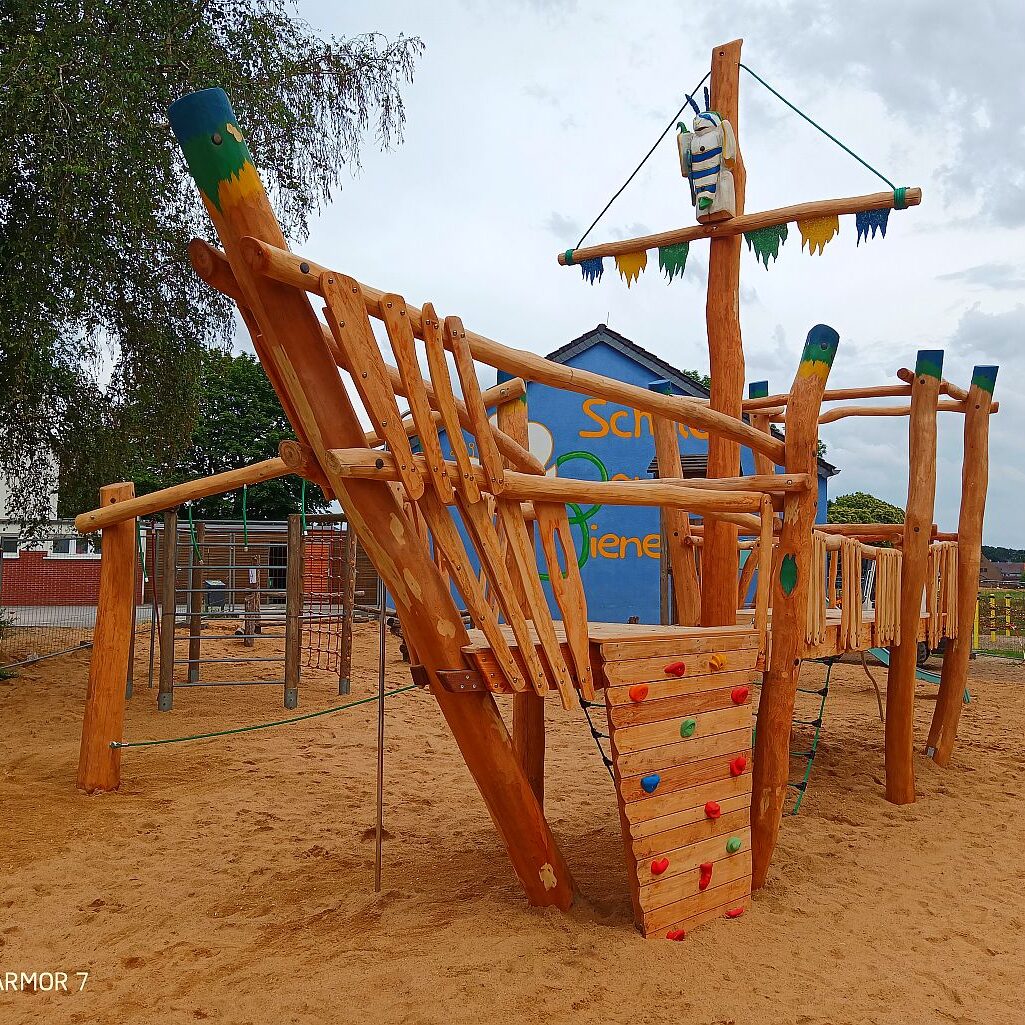 spielschiff-holz-kletterwand-seile-schulhof Holz-Spielschiff auf Sandfläche mit buntem Kletteraufstieg, Masten und Seilen – vor einer blau gestrichenen Schulwand mit Schriftzug