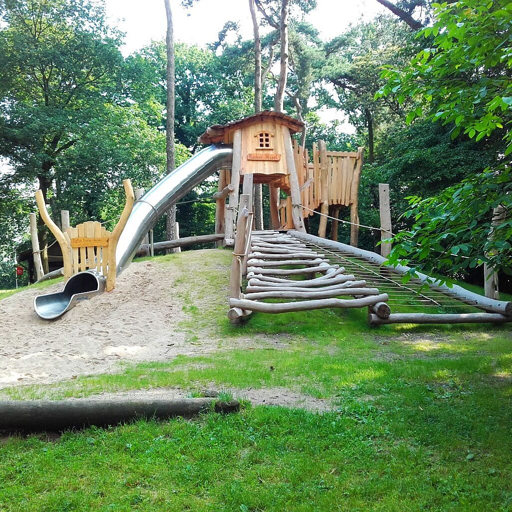waldspielplatz-holzhaus-rutsche-kletternetz Naturnaher Spielplatz mit Holz-Kletterhaus, großer Rutsche, Kletterrampe und Sandfläche – eingebettet im Waldgebiet